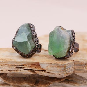 May include: Two rings with green fluorite stones set in copper. The stones are rough and have a natural, irregular shape. The rings are adjustable and have a rustic, bohemian style.