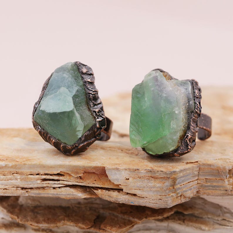 May include: Two rings with green fluorite stones set in copper. The stones are rough and have a natural, irregular shape. The rings are adjustable and have a rustic, bohemian style.