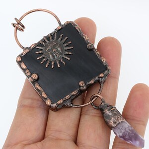 May include: A copper-toned pendant with a black square stone featuring a sun face design. A purple amethyst crystal dangles from the bottom of the pendant.