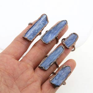 Op de afbeelding: Vijf ringen gemaakt van koperdraad en blauwe kyanite stenen. De ringen zijn in verschillende maten en worden getoond op een hand.