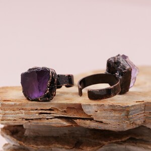 May include: Two adjustable rings with purple amethyst stones set in copper. The rings are on a stacked piece of wood.