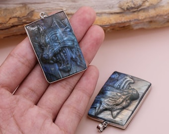 Puede incluir: Dos colgantes de plata con un lobo tallado en una piedra de labradorita azul grisácea. Los colgantes son rectangulares y tienen una anilla de plata.