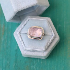 May include: A silver ring with a light pink rectangular gemstone set in a bezel setting. The ring is displayed in a light blue velvet ring box.
