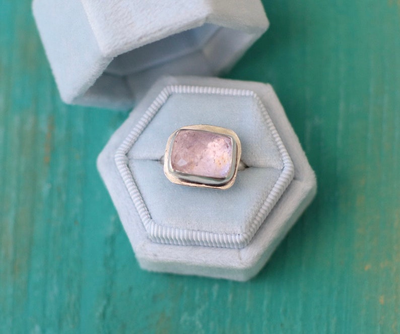 May include: A silver ring with a light pink rectangular gemstone set in a bezel setting. The ring is displayed in a light blue velvet ring box.