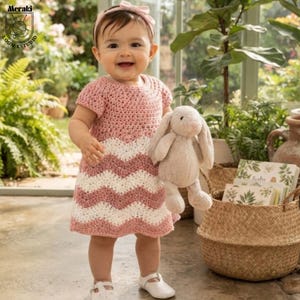 May include: A crochet dress featuring a dusty pink and white chevron pattern with short sleeves and a matching pink headband. The dress is worn with white shoes and is paired with a plush beige rabbit toy held in one hand.