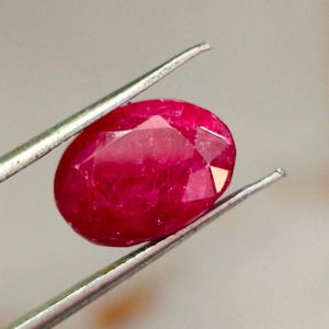 May include: A faceted, oval-shaped ruby gemstone held by silver tweezers. The ruby is a deep, vibrant red colour, with multiple facets catching the light. The tweezers are thin and metallic, providing a secure grip on the gemstone.