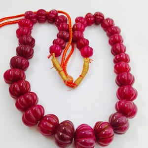 May include: A necklace featuring a series of carved, deep red beads. The beads have a ribbed, pumpkin-like shape and are strung on a red cord. The necklace is displayed against a white background.