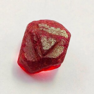 May include: A rough-cut, deep red gemstone with a crystalline structure. The surface has a textured appearance with areas of lighter, metallic-like inclusions. The gem is set against a plain white background, highlighting its natural beauty.