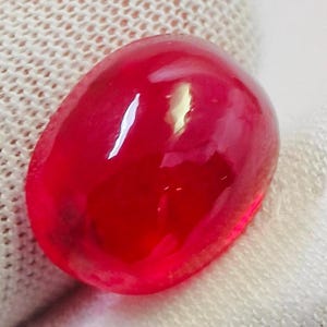 Pode incluir: Uma gema vermelha polida em forma oval. A gema tem uma superfície lisa e brilhante com reflexos internos, sobre um fundo branco texturizado. A cor da pedra é um vermelho profundo e saturado.