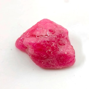 Puede incluir: Una gema en bruto, sin cortar, con un tono rosa intenso y vibrante. La piedra tiene una textura rugosa e irregular y un aspecto ligeramente translúcido. La gema se encuentra sobre un fondo blanco liso.