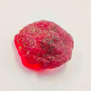 May include: A rough, uncut ruby gemstone. The ruby is a deep red color with a slightly uneven surface. The gemstone has a natural, unpolished appearance, showcasing its raw form. The ruby is a vibrant, eye-catching piece.