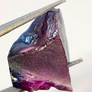 Puede incluir: Una gema en bruto, sin cortar, con un degradado de colores. La piedra transita de un morado intenso en la base a un morado más claro y azul en la parte superior. La gema es sostenida por unas pinzas plateadas, mostrando su textura natural.