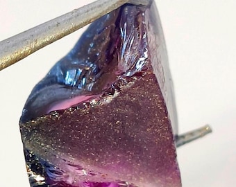 May include: A raw, uncut gemstone with a gradient of colors. The stone transitions from a deep purple at the base to a lighter purple and blue at the top. The gem is held by a pair of silver tweezers, showcasing its rough, natural texture.