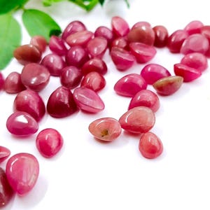 May include: A collection of polished, oval-shaped gemstones in shades of pink and red, scattered on a white surface. The gemstones vary in size and have a smooth, glossy texture. Green leaves are visible in the background.