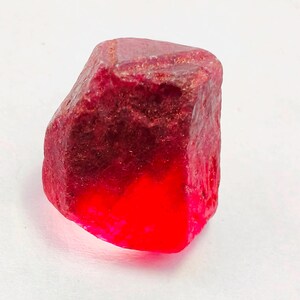 Könnte beinhalten: Ein roher, natürlicher Rubin-Edelstein. Der Rubin hat eine tiefe, gesättigte rote Farbe mit einer leicht unebenen Oberfläche. Der Boden des Steins hat einen hellen, leuchtenden roten Farbton. Der Edelstein ist vor einem weißen Hintergrund platziert.