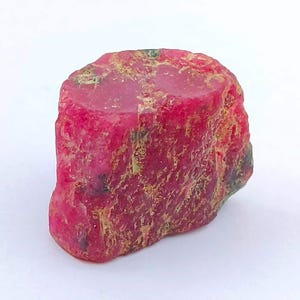 May include: A rough-cut ruby gemstone with a vibrant red hue. The natural stone displays a textured surface with hints of green and gold. This raw ruby is a striking example of natural mineral beauty.
