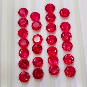 May include: A collection of round, faceted, bright red gemstones arranged in neat rows. The gems have a polished, reflective surface, showcasing their vibrant color and clarity. The gemstones are set against a white, ridged background.