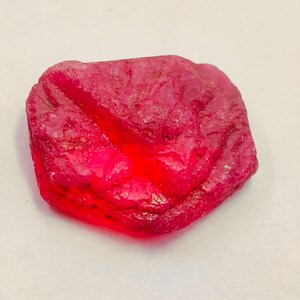 May include: A rough-cut, natural ruby gemstone. The ruby is a deep, saturated red color with a slightly translucent appearance. The surface has a textured, uneven appearance, with visible ridges and variations in color intensity. The gemstone is irregularly shaped.