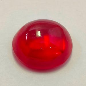 Może przedstawiać: Wypolerowany kamień szlachetny o szlifie kaboszonowym w głębokim, bogatym odcieniu czerwieni. Gładki, zaokrąglony kształt odbija światło, tworząc refleksy i cienie. Kamień szlachetny jest umieszczony na neutralnym tle.