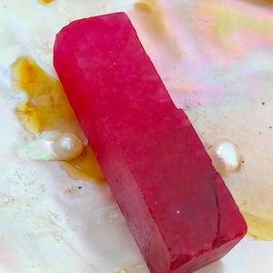 May include: A rectangular, deep red gemstone with a faceted surface. The gemstone is set on a pearlescent, iridescent surface with a pearl. The gemstone's color is a vibrant, saturated red.