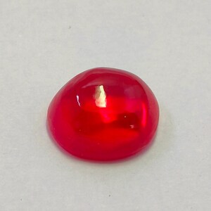 Op de afbeelding: Een glanzende, rode cabochon edelsteen. De gladde, afgeronde vorm reflecteert licht en creëert highlights. De edelsteen is geplaatst tegen een neutrale achtergrond.