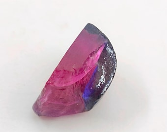 May include: A cut gemstone with a vibrant color gradient. The stone transitions from a deep magenta to a dark, almost black, hue. Hints of blue and pink are visible within the translucent material. The shape is a partial wedge.