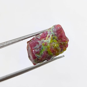 May include: A rough-cut gemstone held by tweezers. The stone displays a mix of colors, including red, green, and yellow. The surface of the stone has a textured appearance, with a slightly irregular shape. The background is plain white.