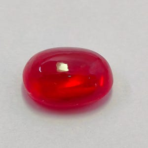 Op de afbeelding: Een gladde, ovaalvormige, rode edelsteen. De edelsteen heeft een glanzend, reflecterend oppervlak, met een kleine kleurvariatie. De steen is geplaatst tegen een effen, gebroken witte achtergrond. De edelsteen is een cabochon-slijpsel.