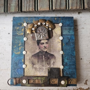 May include: Mixed media artwork featuring a vintage portrait of a woman wearing a decorative headpiece. The artwork is mounted on a blue and gold patterned background, with metal and white embellishments. The piece includes a metal frame with the numbers 3, 5, and 7.
