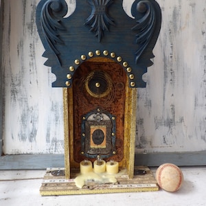 May include: A blue and gold wooden shrine with a small picture inside, three white candles, and a baseball on the base.
