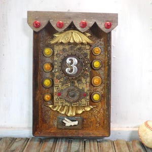 May include: A rustic, mixed-media wall art piece with a weathered, brown metal base. It features a prominent silver number "3" surrounded by gears, gold accents, and colorful embellishments. A baseball rests nearby.
