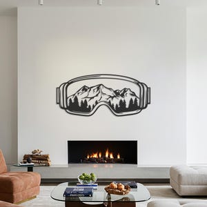 Könnte beinhalten: Schwarze Metall-Wandkunst, die eine Skibrille mit einer Berg- und Waldszene im Inneren darstellt. Das Kunstwerk ist an einer weißen Wand über einem Kamin angebracht.