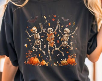 Puede incluir: Camiseta negra con un diseño de Halloween. El gráfico muestra tres esqueletos bailando rodeados de hojas de otoño y calabazas. Dos murciélagos vuelan por encima. Los esqueletos tienen los brazos levantados en una pose alegre. Las calabazas son naranjas.