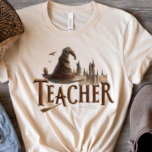 May include: Cream-colored t-shirt featuring a brown wizard hat, owl, and castle graphic. The word "TEACHER" is printed in brown, with a broom below. A casual, comfortable tee for educators.