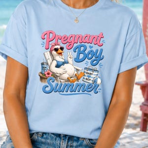 May include: Pale blue t-shirt featuring a cartoon goose in sunglasses and a pink headband, giving a peace sign. The text says "Pregnant Boy Summer" in pink and blue. A summer-themed design.