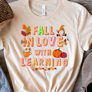 Puede incluir: Camiseta beige con el texto "FALL IN LOVE WITH LEARNING" en letras coloridas. El diseño incluye gráficos temáticos de otoño como calabazas, hojas, un gnomo y una taza de café.