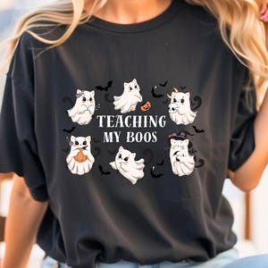 May include: Black t-shirt featuring a Halloween-themed design. The graphic includes cartoon ghosts with various accessories, bats, and the text "TEACHING MY BOOS" in white. The ghosts are white with black eyes and are holding pumpkins and wearing hats.