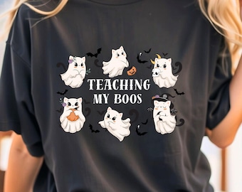 May include: Black t-shirt with a Halloween design. The graphic shows cartoon ghosts with accessories, bats, and the text "TEACHING MY BOOS" in white. The ghosts are white with black eyes and are holding pumpkins and wearing hats.