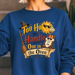 May include: Blue sweatshirt with a graphic that reads "Too Hot to Handle, One in the Oven." The design includes a witch hat, broom, sunglasses-wearing sun, and a baby-sized beach chair with a onesie that says "Coming Soon."