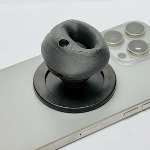 May include: A black phone grip with a round base and a textured, spherical top. The grip is attached to a silver smartphone.