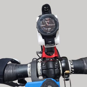 May include: A black Garmin smartwatch attached to a bicycle handlebar. The watch shows the date, time, and battery level. The handlebar is black with red and white details, and the mount is white.