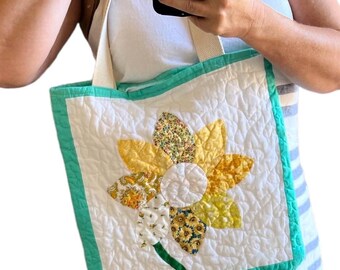 May include: A white quilted tote bag featuring a large, multi-patterned yellow and gold flower appliqué on the front. The bag has a bright teal border and two cream-colored fabric handles. The quilted texture covers the entire white surface.