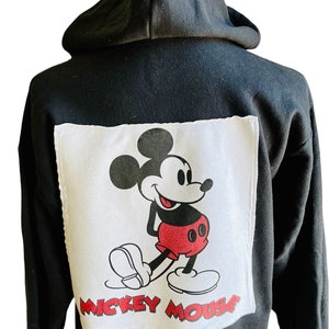 May include: Black hooded sweatshirt with a white square patch featuring a cartoon image of Mickey Mouse in red shorts and black shoes. The text "Mickey Mouse" is printed below the image.