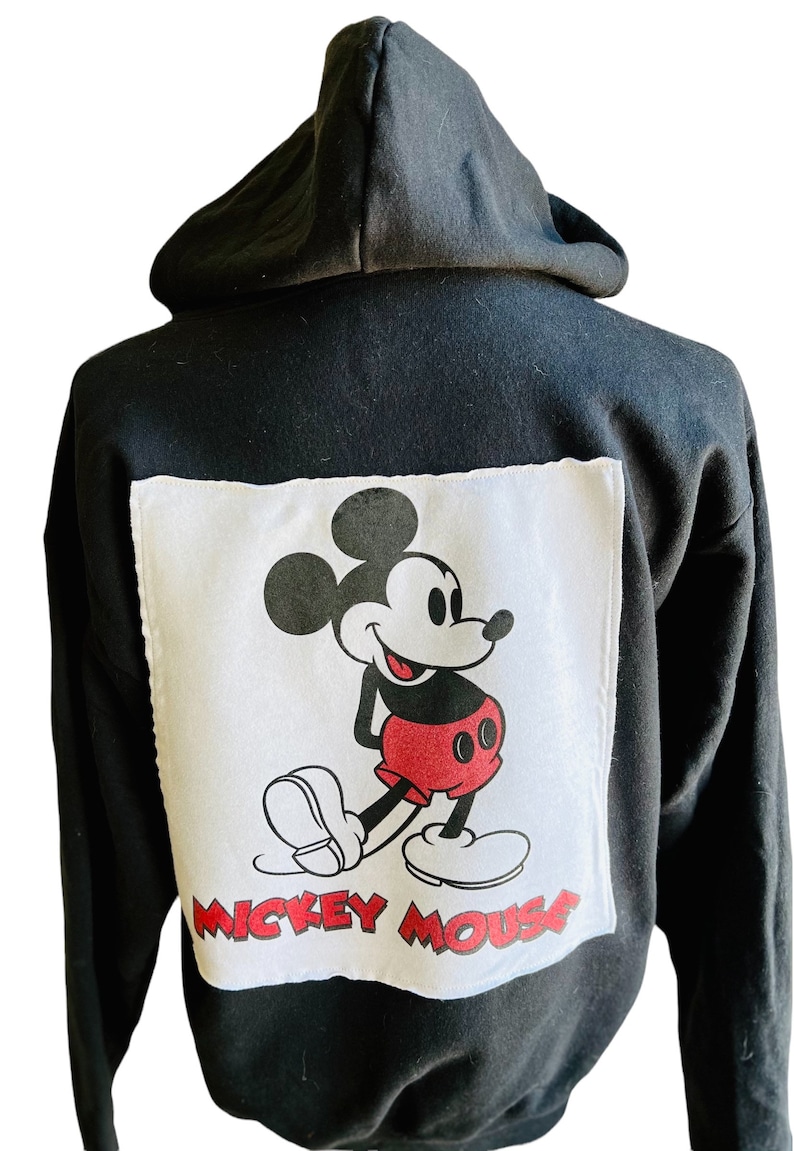 May include: Black hooded sweatshirt with a white square patch featuring a cartoon image of Mickey Mouse in red shorts and black shoes. The text "Mickey Mouse" is printed below the image.