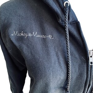 May include: Black zip-up hoodie with a white embroidered design that says "Mickey Mouse" with a heart symbol between the words.
