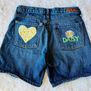 May include: Blue denim shorts with a floral heart patch on the back left pocket and a daisy with the word "DAISY" embroidered on the back right pocket.