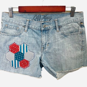 May include: Light blue denim shorts with a frayed hem and a patchwork flower design in red, white, and blue on the front left leg. The shorts have a brand label that reads "Old Navy".