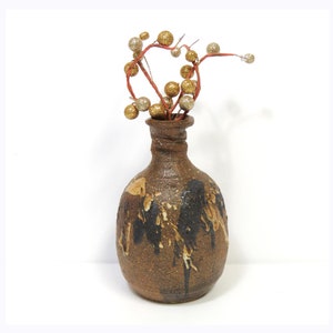 May include: A small, brown ceramic vase with a textured surface and black and white paint accents. The vase is filled with a sprig of red twigs with gold and silver glitter balls.