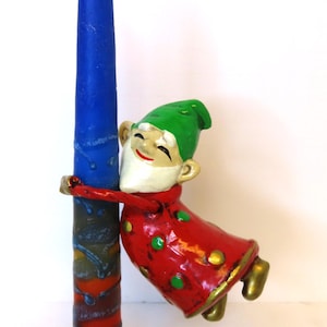 May include: A tall, cylindrical blue candle with a textured, multi-colored base. A small gnome figurine with a green hat, white beard, and red coat with green polka dots is attached to the side of the candle, appearing to hug it tightly.
