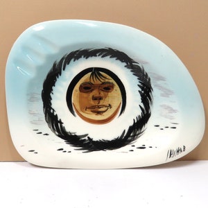 May include: A vintage ashtray with a light blue and white design. The center features a face with a black, circular border. The face is brown with black hair and a smiling expression. The ashtray has an irregular shape.